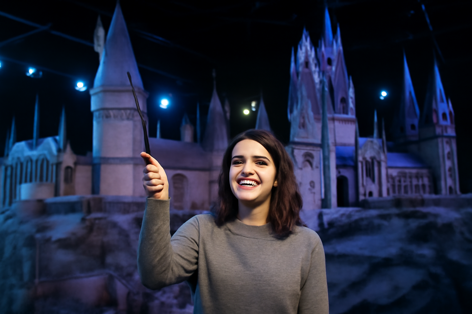 Visitor smiling with excitement while waving a wand in front of the intricately detailed Hogwarts Castle model at the Harry Potter Studio Tour London. The magical atmosphere is enhanced by cool lighting and the castle's towering spires.